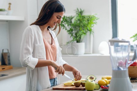 Téléchargez les photos : Tourné de beauté femme coupe pepino pour préparer un jus de désintoxication dans la cuisine à la maison. - en image libre de droit