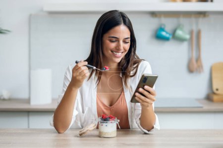 Tiro de hermosa mujer comiendo una papilla con bayas mientras usa el teléfono inteligente en la cocina en casa