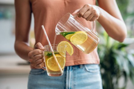 Téléchargez les photos : Tourné de jeune femme tenant et servant un jus de désintoxication dans la cuisine à la maison - en image libre de droit