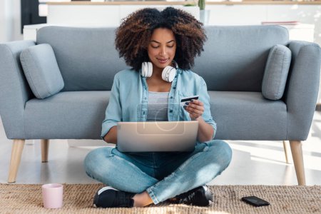 Tiro de mujer relajante que paga algo en línea con su tarjeta de crédito con el ordenador portátil mientras está sentado en el suelo en la sala de estar en casa
