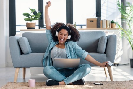 Foto de Foto de mujer motivada que paga algo en línea con su tarjeta de crédito con el ordenador portátil mientras celebra sentado en el suelo en la sala de estar en casa - Imagen libre de derechos
