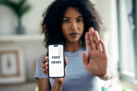 Tiro de mujer seria sosteniendo el teléfono móvil con el mensaje de noticias falsas mientras hace señal de stop con la mano en casa