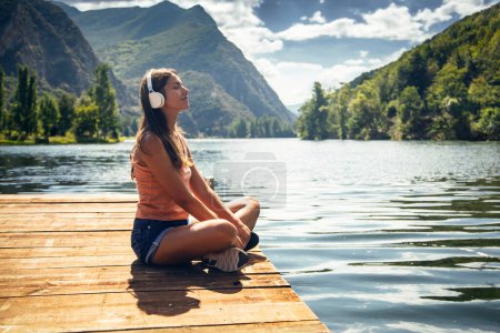 Foto de Foto de mujer feliz escuchando música con auriculares mientras se relaja sentado junto a un lago - Imagen libre de derechos