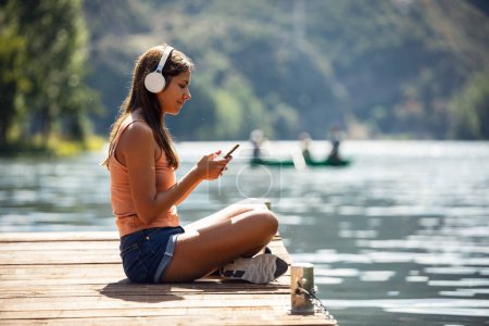 Téléchargez les photos : Prise de vue de belle femme écoutant de la musique avec des écouteurs tout en utilisant un smartphone près du lac - en image libre de droit