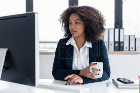 Foto de Tiro de mujer de negocios elegante concentrado que trabaja con el ordenador mientras bebe café en una oficina moderna - Imagen libre de derechos