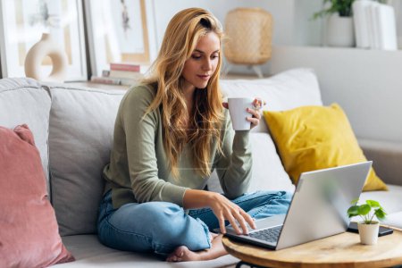 Téléchargez les photos : Prise de vue de jeune femme confiante travaillant avec son ordinateur portable tout en buvant une tasse de café assis sur un canapé à la maison - en image libre de droit