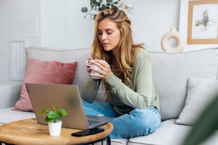 Téléchargez les photos : Prise de vue d'une jeune femme joyeuse travaillant avec son ordinateur portable tout en buvant une tasse de café assis sur un canapé à la maison - en image libre de droit