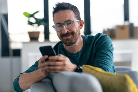 Photo for Shot of happy mature man with eyeglasses using his smartphone while relaxing in the couch at home. - Royalty Free Image