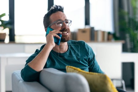 Foto de Tiro de hombre maduro feliz con anteojos hablando con su teléfono inteligente mientras se relaja en el sofá en casa. - Imagen libre de derechos