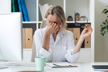 Téléchargez les photos : Prise de vue du médecin femme mature ayant mal à la tête tout en travaillant avec l'ordinateur dans la consultation. - en image libre de droit