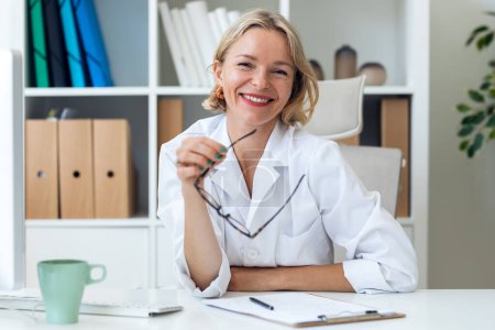 Téléchargez les photos : Prise de vue d'une femme mûre souriante médecin souriant regardant la caméra tout en travaillant avec un ordinateur portable dans la consultation - en image libre de droit