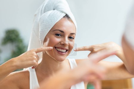 Téléchargez les photos : Portrait de belle jeune femme prenant soin de sa peau tout en mettant de la crème en regardant miroir à la maison. - en image libre de droit
