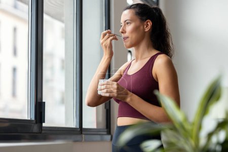 Téléchargez les photos : Tourné de belle femme sportive mangeant du yaourt tout en regardant vers l'avant par la fenêtre à la maison. - en image libre de droit