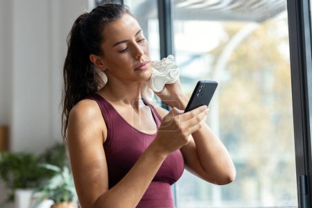 Téléchargez les photos : Prise de vue de femme sportive utilisant un téléphone portable tout en essuyant la sueur avec une serviette après l'entraînement à la maison - en image libre de droit