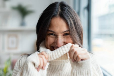 Téléchargez les photos : Tourné de mignonne femme souriante couvrant son visage avec un pull en laine blanche à la maison - en image libre de droit