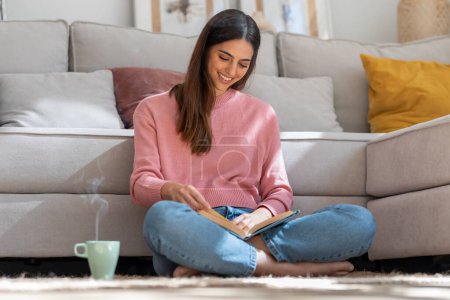 Téléchargez les photos : Prise de vue d'une jolie jeune femme lisant un livre et buvant une tasse de café alors qu'elle était assise par terre à la maison. - en image libre de droit