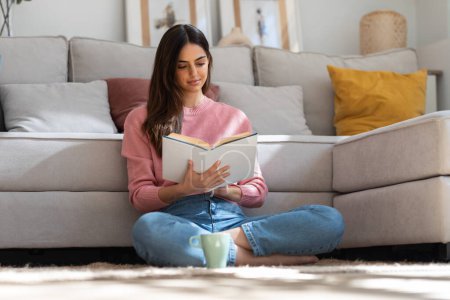 Foto de Tiro de mujer joven y bonita leyendo un libro y bebiendo una taza de café mientras está sentada en el suelo en casa. - Imagen libre de derechos