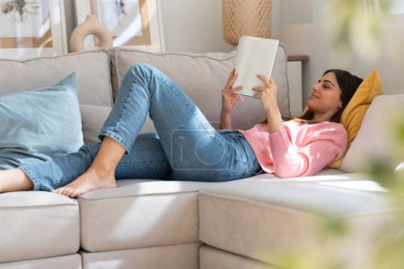 Téléchargez les photos : Prise de vue de jolie jeune femme lisant un livre alors qu'elle était allongée sur un canapé à la maison - en image libre de droit