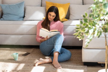 Photo for Shot of pretty young woman reading a book and drinking a cup of coffee while sitting on the floor at home. - Royalty Free Image
