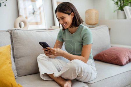 Téléchargez les photos : Prise de vue d'une jeune femme souriante utilisant son téléphone portable alors qu'elle était assise sur un canapé à la maison - en image libre de droit