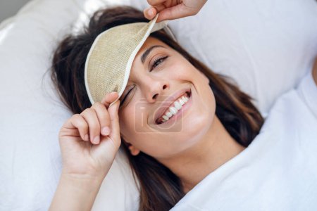 Téléchargez les photos : Portrait de jolie jeune femme tirant vers le haut masque de sommeil et regardant la caméra après le réveil dans la chambre à coucher à la maison
. - en image libre de droit