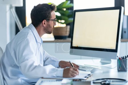 Téléchargez les photos : Prise de vue du médecin masculin mature travaillant avec l'ordinateur tout en écrivant des notes dans la consultation médicale - en image libre de droit