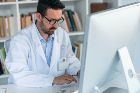 Téléchargez les photos : Prise de vue du médecin masculin mature travaillant avec l'ordinateur tout en écrivant des notes dans la consultation médicale - en image libre de droit