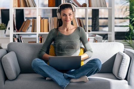 Foto de Foto de una hermosa mujer joven que trabaja con su computadora portátil mientras escucha música con auriculares sentados en un sofá en casa. - Imagen libre de derechos