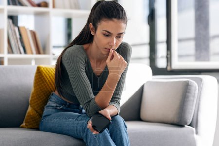 Foto de Tiro de mujer joven deprimida pensando en sus problemas mientras sostiene el teléfono móvil sentado en el sofá en casa. - Imagen libre de derechos