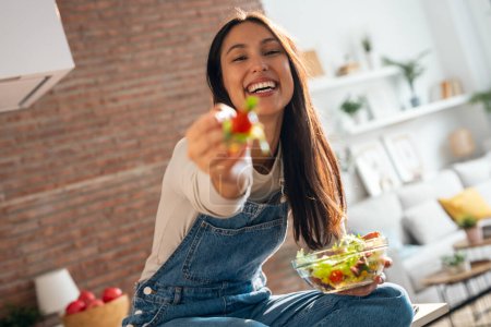 Téléchargez les photos : Portrait d'une belle femme souriante mangeant une salade saine assis sur la table de la cuisine à la maison. - en image libre de droit