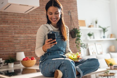 Téléchargez les photos : Tourné de belle jeune femme mangeant un bol de salade tout en utilisant son téléphone portable dans la cuisine à la maison. - en image libre de droit