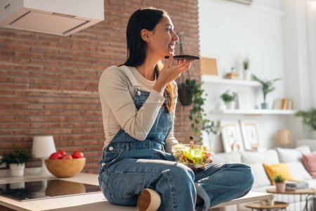 Téléchargez les photos : Tourné de beauté jeune femme mangeant un bol de salade tout en utilisant son téléphone portable dans la cuisine à la maison. - en image libre de droit