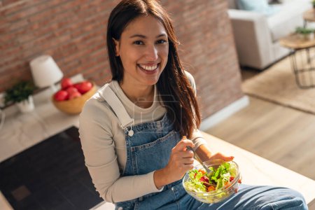 Foto de Retrato de la mujer sonriente belleza comiendo ensalada saludable mientras se sienta en la mesa de la cocina en casa. - Imagen libre de derechos