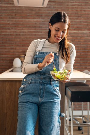 Photo for Shot of pretty smiling woman eating healthy salad standing in the kitchen. - Royalty Free Image