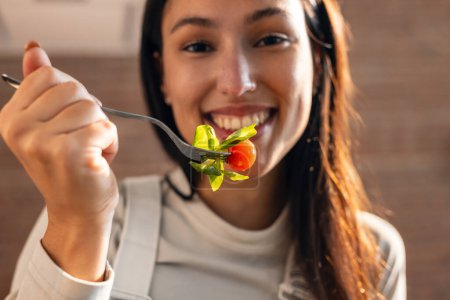 Téléchargez les photos : Gros plan d'une jolie femme souriante mangeant une salade saine assis sur la table de la cuisine à la maison. - en image libre de droit