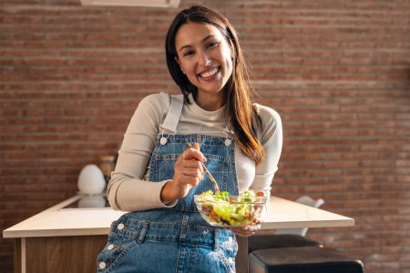 Téléchargez les photos : Portrait de jolie femme souriante mangeant une salade saine tout en regardant la caméra debout dans la cuisine. - en image libre de droit