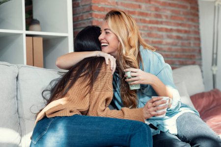 Téléchargez les photos : Prise de vue de deux belles femmes souriantes s'embrassant tout en parlant assis sur le canapé dans le salon à la maison. - en image libre de droit