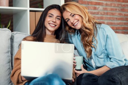 Téléchargez les photos : Shot of two happy beautiful women using computer while planning their vacation sitting on the couch at home. - en image libre de droit