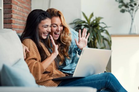 Téléchargez les photos : Prise de vue de deux belles femmes heureuses faisant un appel vidéo avec ordinateur portable tout en agitant la main assise sur le canapé à la maison. - en image libre de droit
