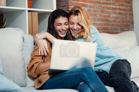 Foto de Shot of two happy beautiful women doing a video call with laptop while hugging each other sitting on the couch at home. - Imagen libre de derechos