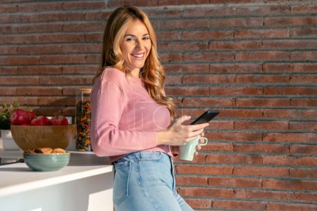 Foto de Tiro de mujer joven sonriente usando su teléfono móvil mientras bebe una taza de café en la cocina en casa. - Imagen libre de derechos