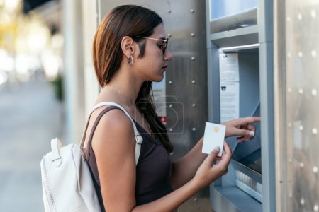 Téléchargez les photos : Prise de vue de belle jeune femme prenant de l'argent du distributeur automatique de billets avec carte de crédit dans la ville. - en image libre de droit