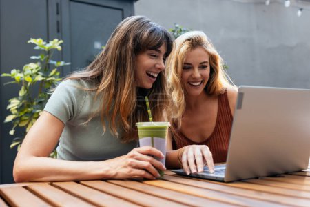 Foto de Foto de dos amigos felices compartiendo un brunch juntos mientras trabajan con el ordenador portátil en la terraza de la cafetería ecológica. - Imagen libre de derechos