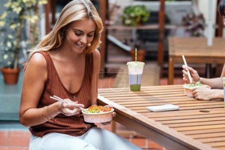 Téléchargez les photos : Tourné de deux belles femmes souriantes amies mangeant des baguettes avec des baguettes tout en parlant sur la terrasse du restaurant. - en image libre de droit