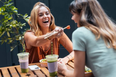 Téléchargez les photos : Tourné de deux belles femmes souriantes amies mangeant des baguettes avec des baguettes tout en parlant sur la terrasse du restaurant. - en image libre de droit
