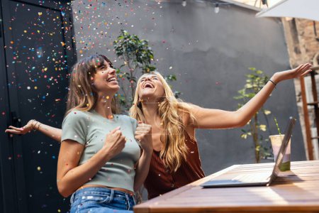 Téléchargez les photos : Tourné de jolies amies mignonnes s'amusant avec des confettis tout en s'enregistrant avec leur ordinateur portable sur une terrasse - en image libre de droit