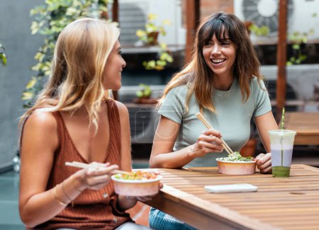 Photo for Shot of two smiling beautiful women friends eating pokes with chopsticks while talking in the restaurant terrace. - Royalty Free Image