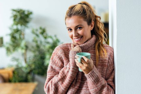 Téléchargez les photos : Tourné de belle jeune femme buvant une tasse de café chaud tout en regardant la caméra dans le salon à la maison. - en image libre de droit