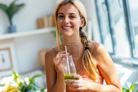 Téléchargez les photos : Portrait de femme heureuse buvant un smoothie vert sain tout en regardant la caméra debout dans le salon à la maison. - en image libre de droit