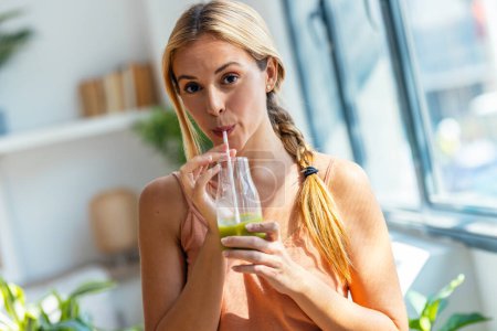 Foto de Retrato de una linda mujer bebiendo un batido verde saludable mientras mira a la cámara de pie en la sala de estar en casa. - Imagen libre de derechos